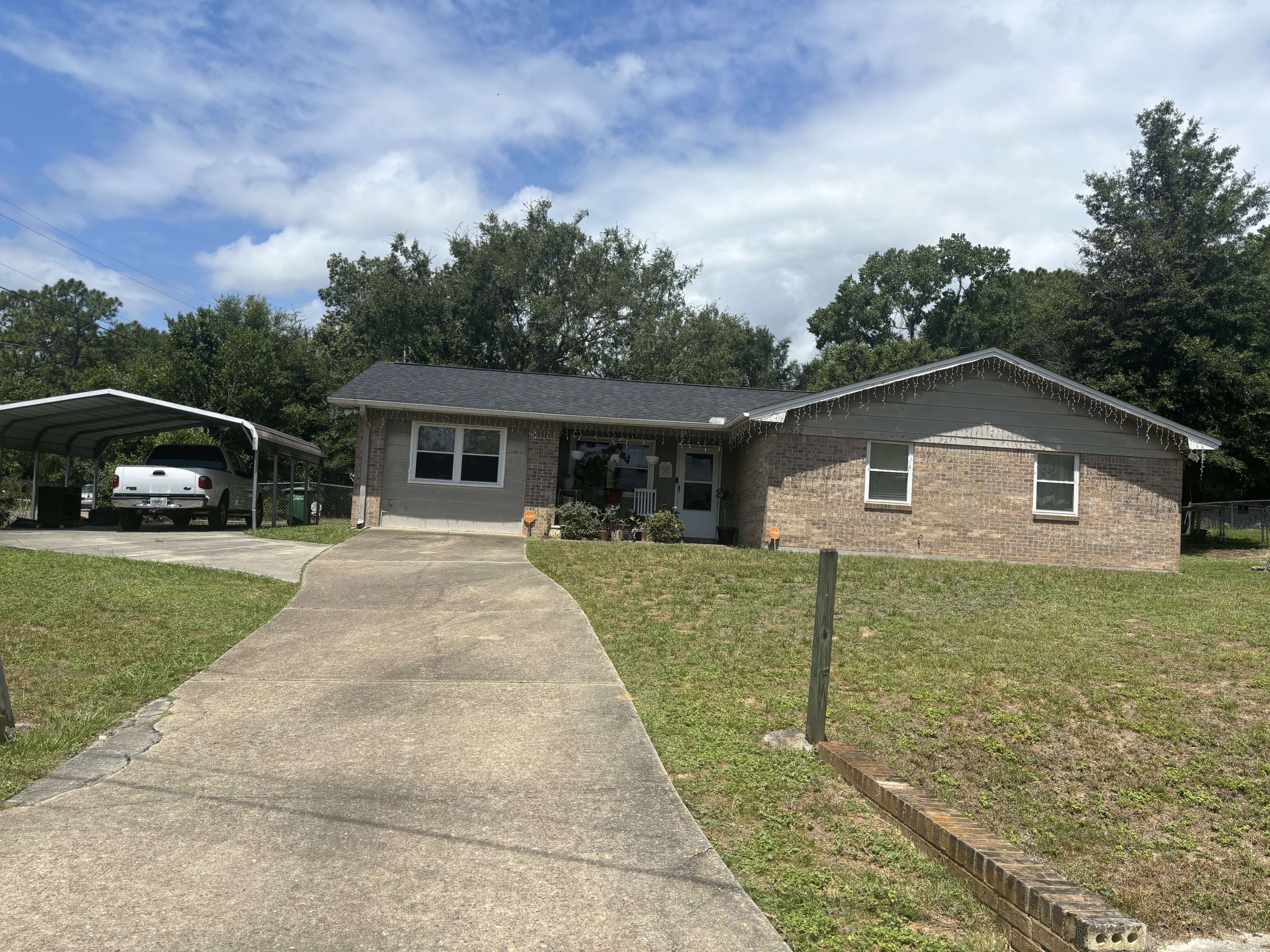 Single-story brick house with a black roof, wide driveway, carport on the left with a white vehicle, and a large front yard with minimal landscaping.