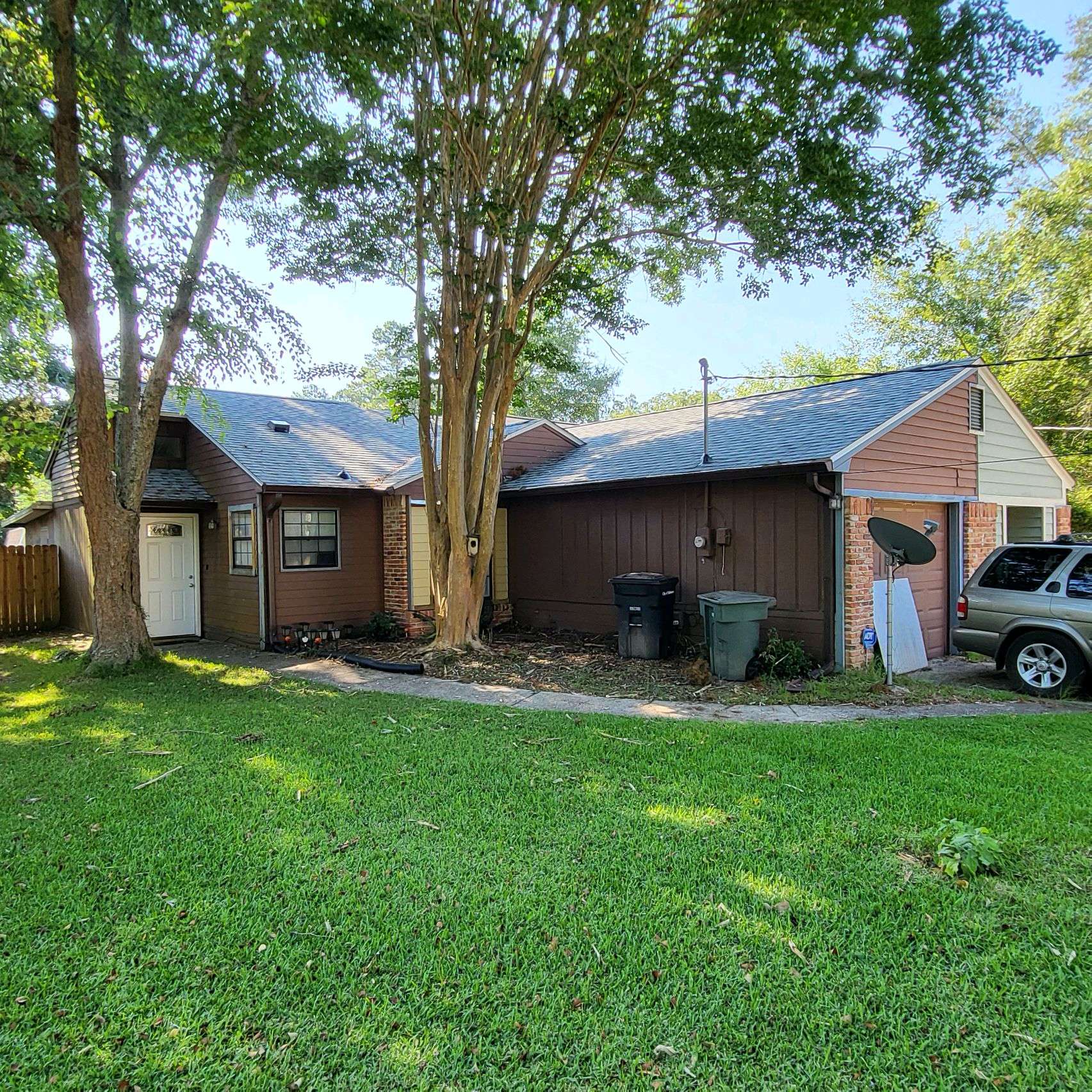 Single-story house with brick and wood siding, two large trees in front yard, trash bins by the wall, and a parked SUV near the driveway on a sunny day.