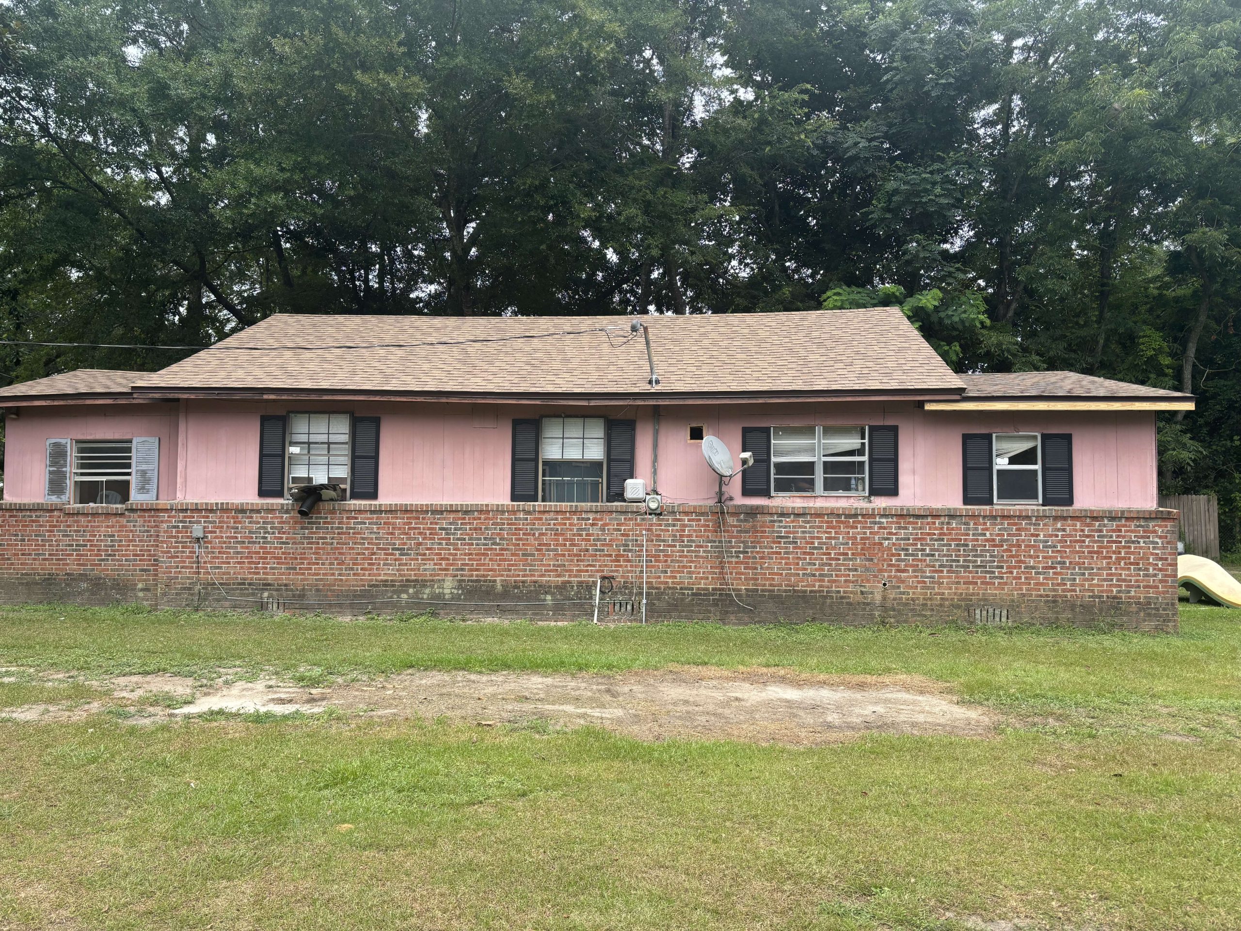Single-story house with a brown shingle roof, pink upper siding, and red brick lower walls. There are black window shutters, a satellite dish, and grass in the foreground.