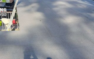 Flat rooftop with fresh roofing material, a single vent pipe, a ladder at the edge, and trees in the background. Shadows and sunlight are visible on the surface.