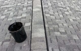 A close-up view of a shingled roof with a ridge vent installation in progress; a black bucket sits on the roof near the work area.