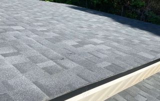 A view of a roof with new, dark gray asphalt shingles under bright sunlight, surrounded by green trees in the background.