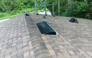 A rooftop with brown asphalt shingles, a black attic vent, tools, gloves, and a satellite dish installation in progress surrounded by green trees.