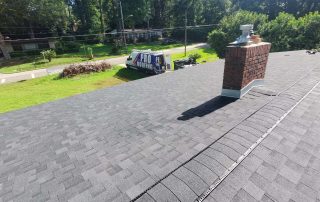 A newly shingled roof with a brick chimney is shown from above; a Pro Roofing truck is parked on the grass near residential homes and trees.