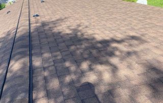 A view looking down a sloped roof with brown asphalt shingles, with tree shadows cast over the surface; houses, grass, and trees are visible in the background.