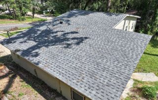 A house with a new gray shingled roof, seen from above, surrounded by trees and a grassy yard.