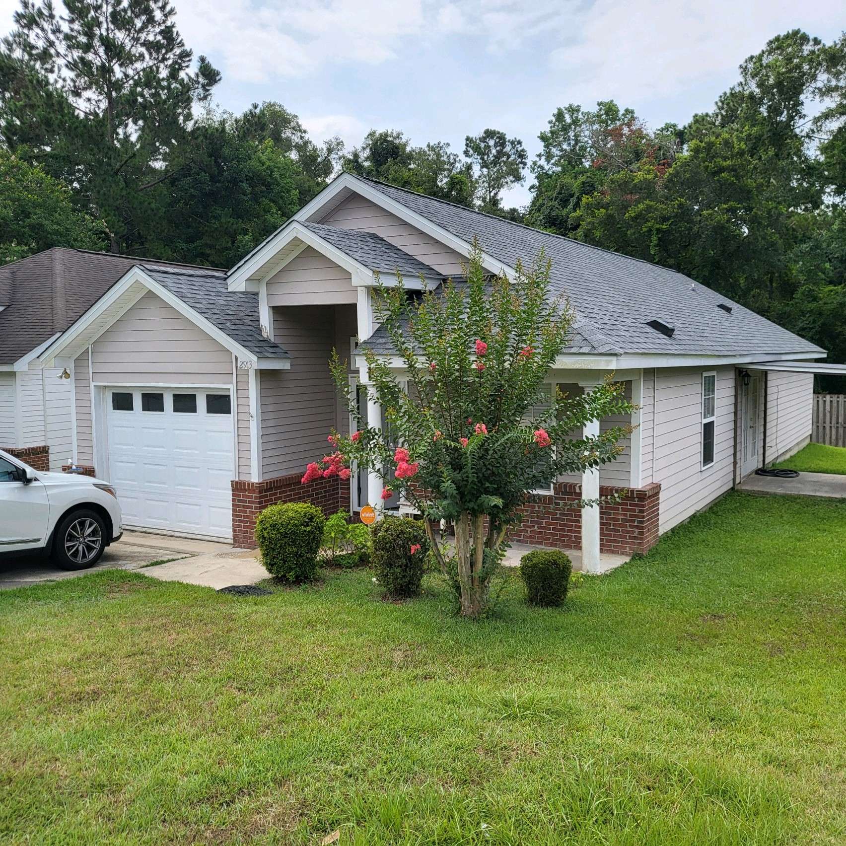 Single-story house with light gray siding, white trim, attached garage, and front yard featuring a small flowering tree and shrubs. A white car is parked in the driveway.