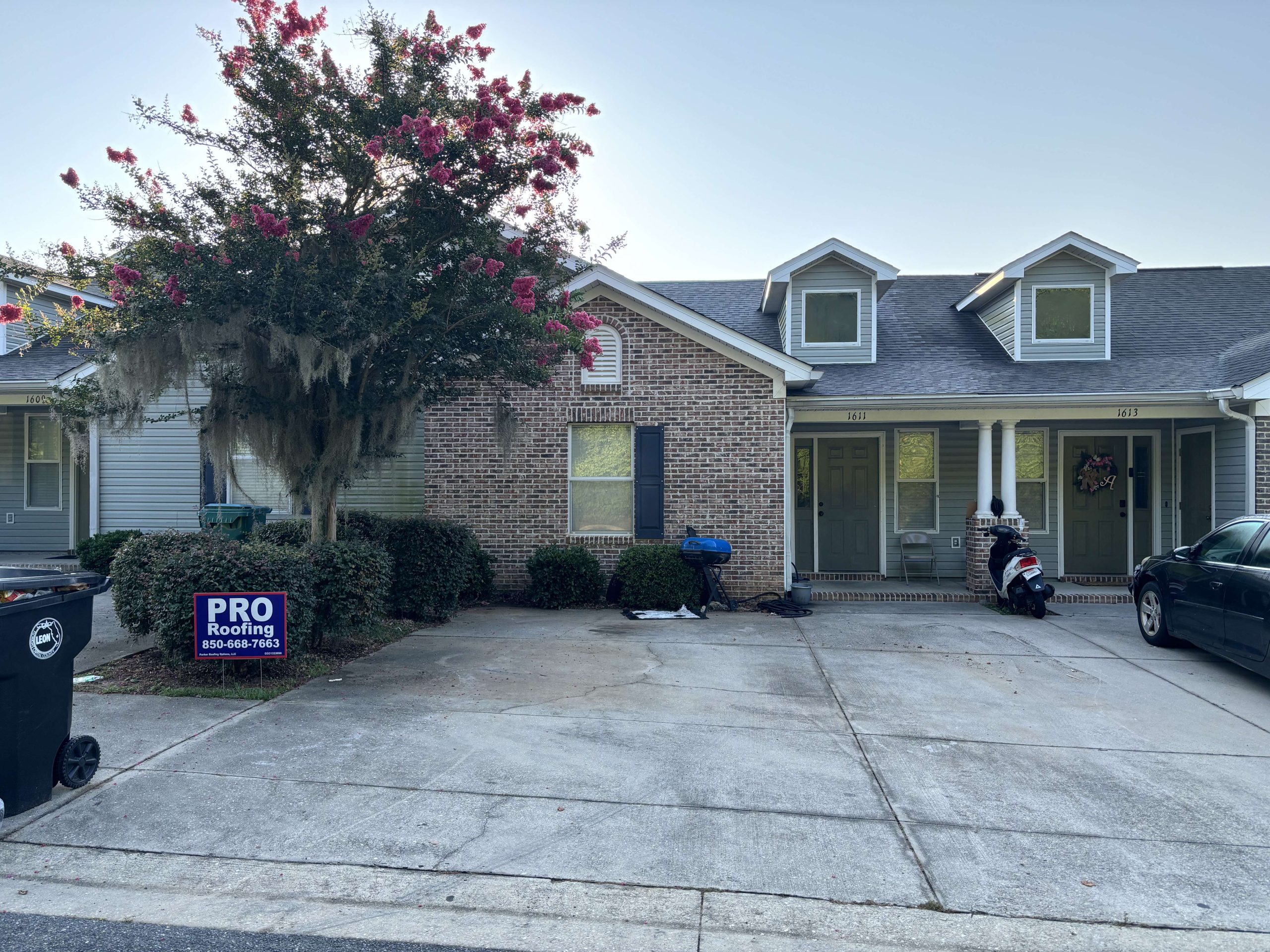 Brick townhouse with two front doors, a flowering tree, a scooter parked by the entrance, and a Pro Roofing sign on the lawn next to a trash bin.