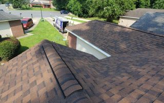 A close-up view of brown shingle roofs on suburban houses, with a lawn, a red car, and a white trailer visible in the background.