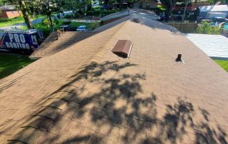 A brown shingle roof viewed from above, with a vent and pipe protruding; a Pro Roofing van is parked beside the house in a residential neighborhood.