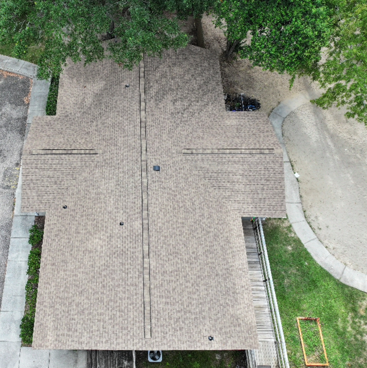 Aerial view of a house with a brown shingle roof surrounded by trees, grass, concrete paths, and a dirt area.
