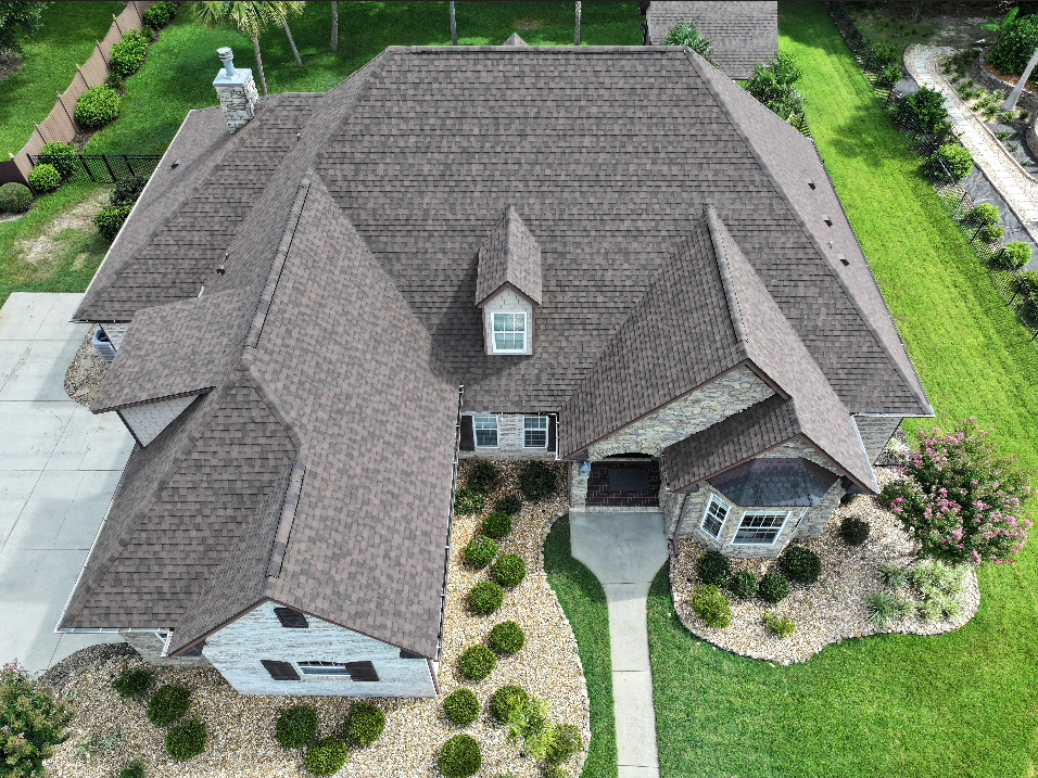 Aerial view of a large house with a brown shingle roof, stone exterior, driveway, walkway, and landscaped yard with bushes and green grass.
