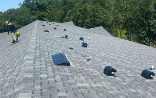 A person with equipment walks on a gray shingled roof with multiple roof vents and scattered tools, surrounded by trees under a clear blue sky.