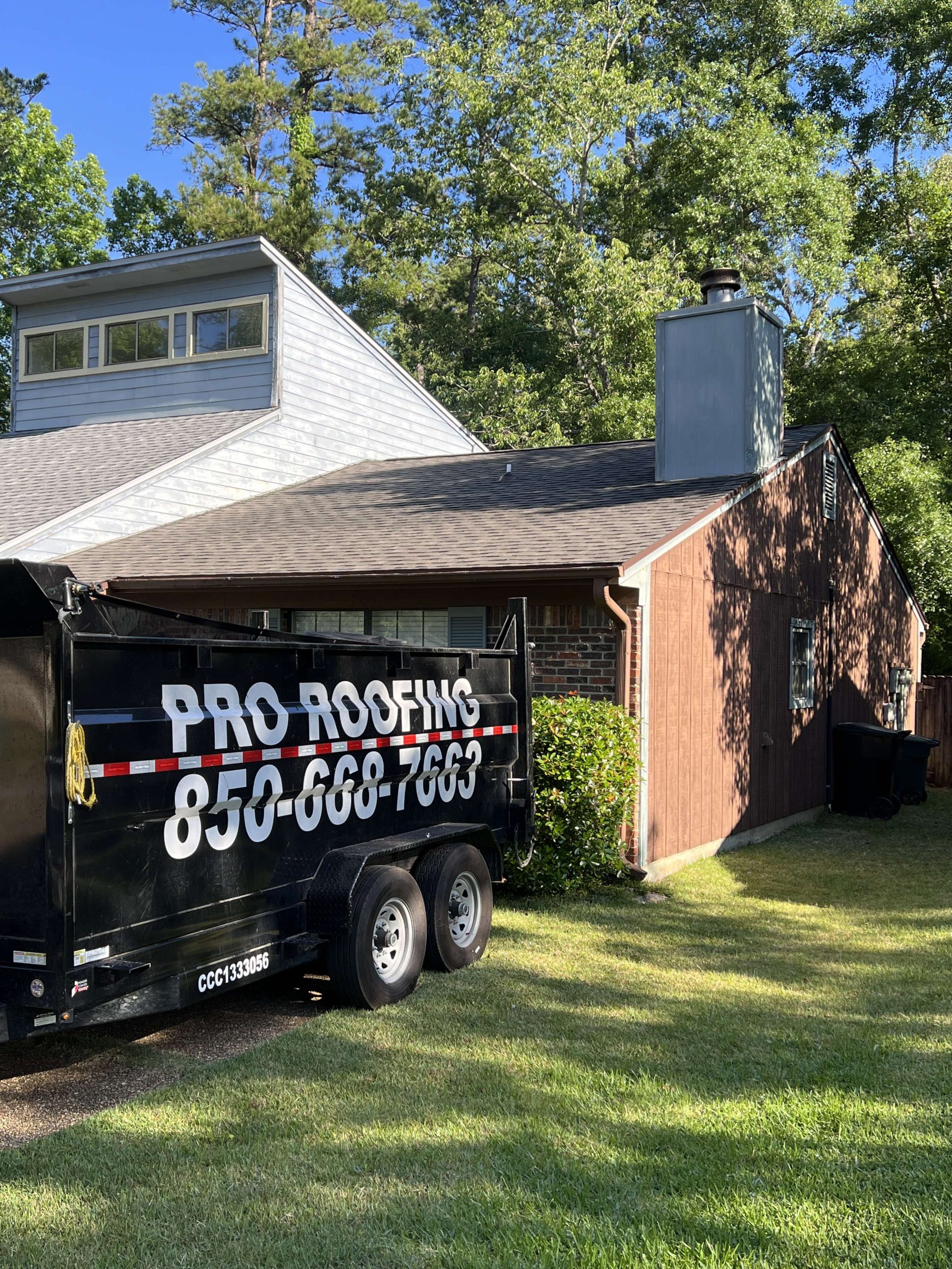 A black trailer labeled PRO ROOFING 850-668-7663 is parked on grass beside a brown house with a chimney, under clear skies.