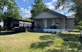 A gray single-story house with a covered porch sits on a grassy lawn. A black Pro Roofing trailer is parked in the driveway beside the house.