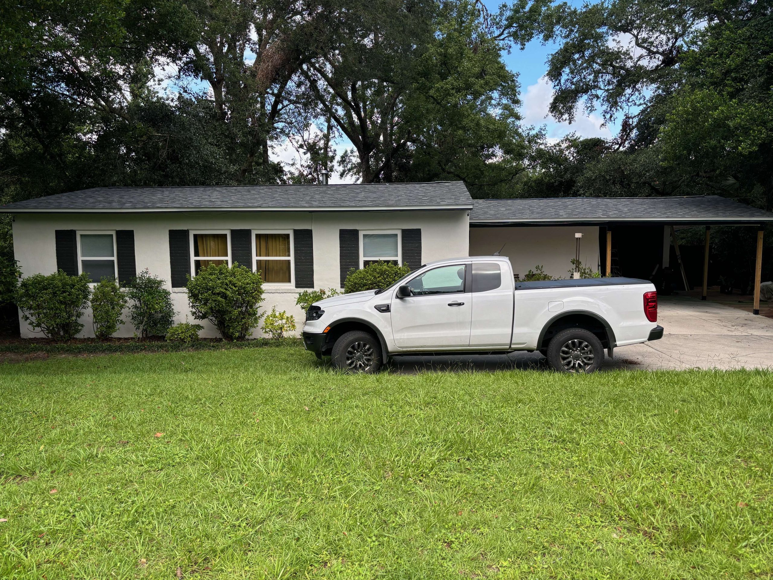 A white pickup truck is parked on the driveway in front of a single-story house with dark shutters and a carport, surrounded by green grass and trees.