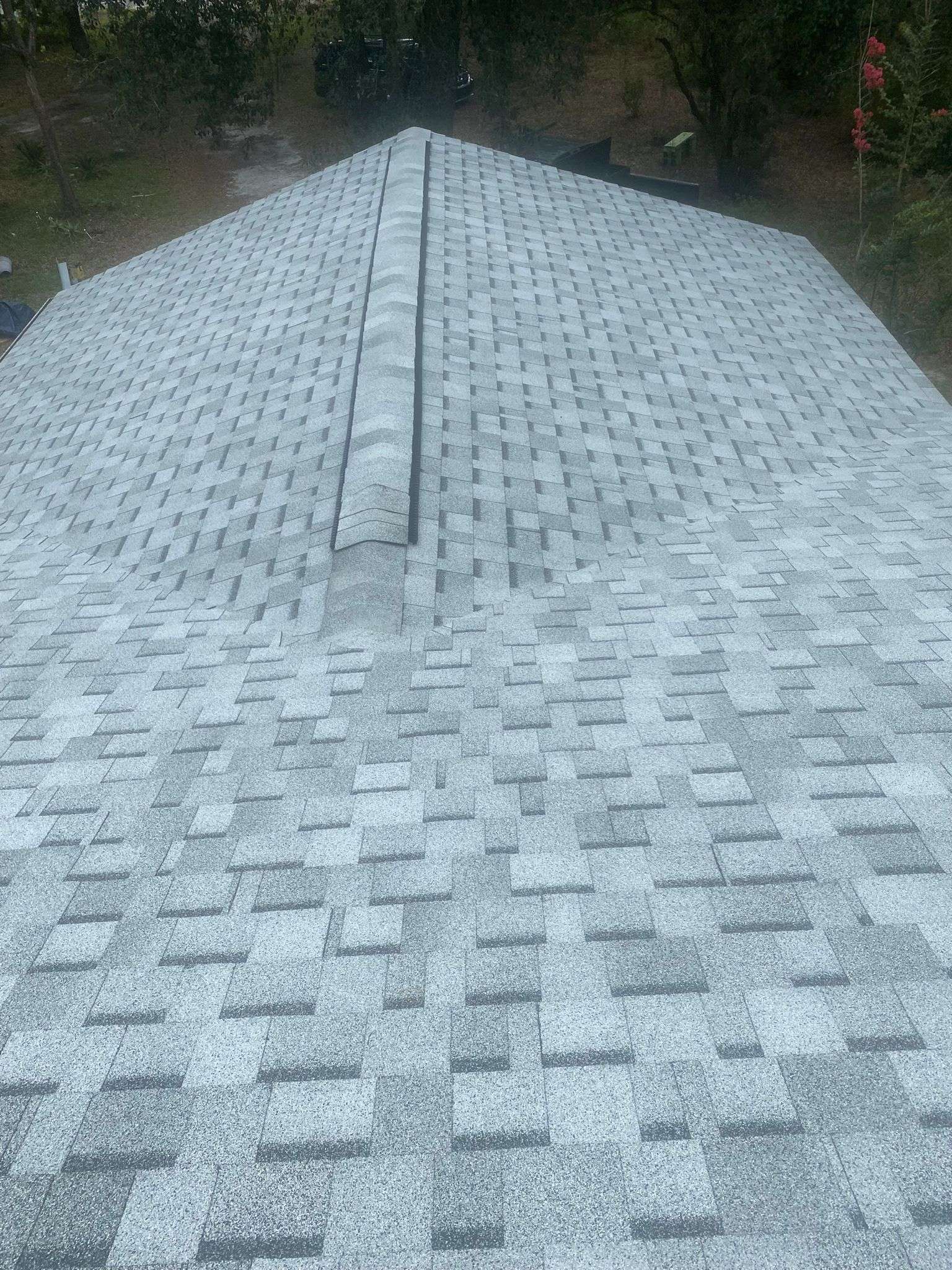 A newly installed gray asphalt shingle roof with a ridge vent, photographed from above, surrounded by trees and a grassy yard.