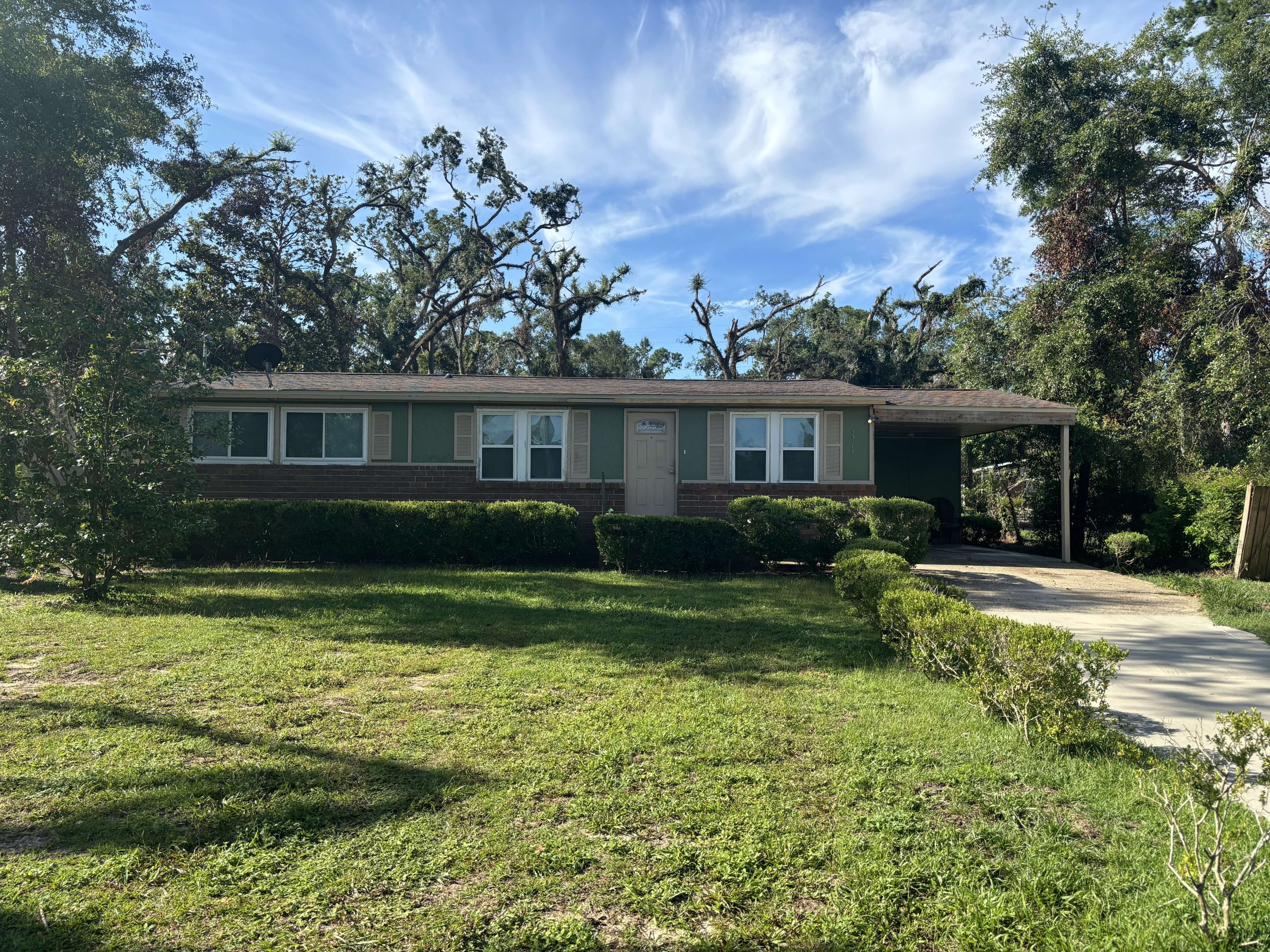 Single-story house with green siding and brick trim, central front door, small bushes in front, carport on right, and a driveway, set against a partly cloudy sky.