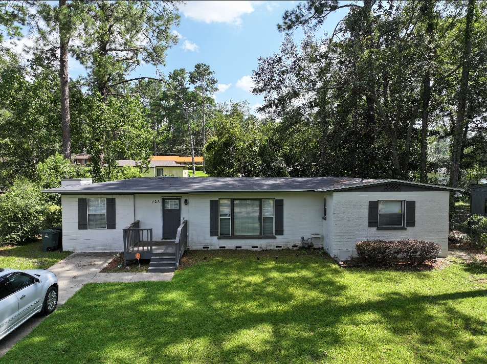 Single-story white brick house with dark shutters, large front windows, a small porch, and a grassy lawn, surrounded by trees. A white car is parked in the driveway.