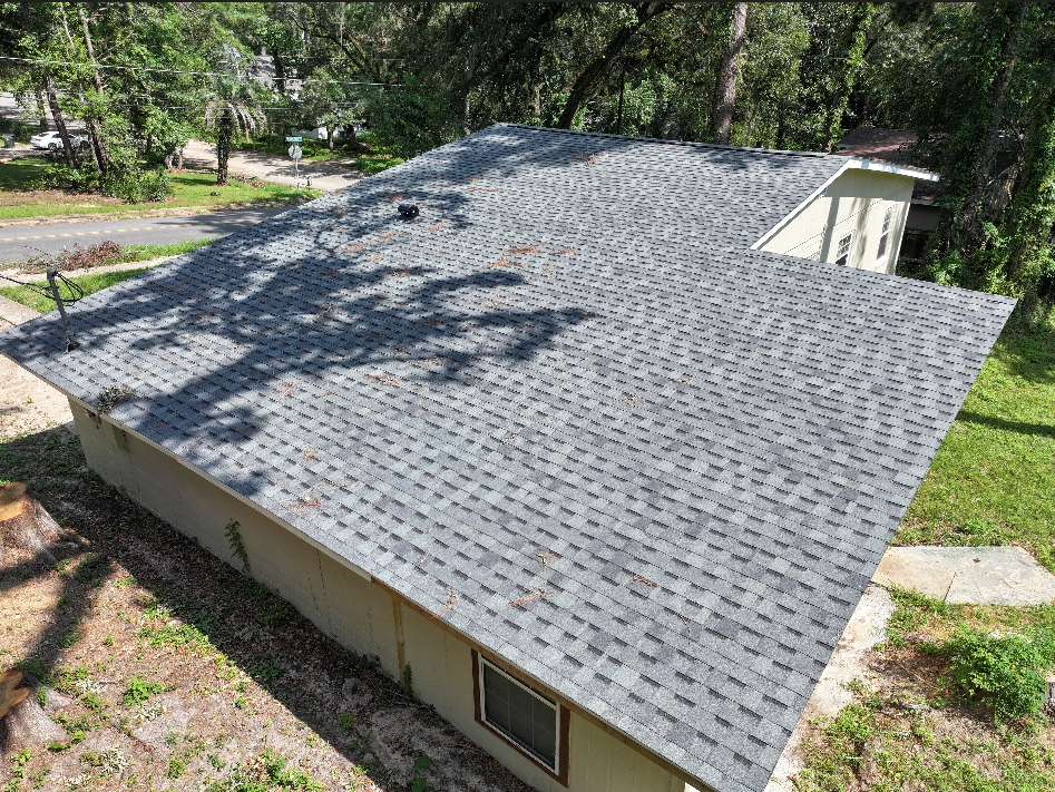 A house with a gray asphalt shingle roof, surrounded by trees and grass, viewed from above during the daytime.