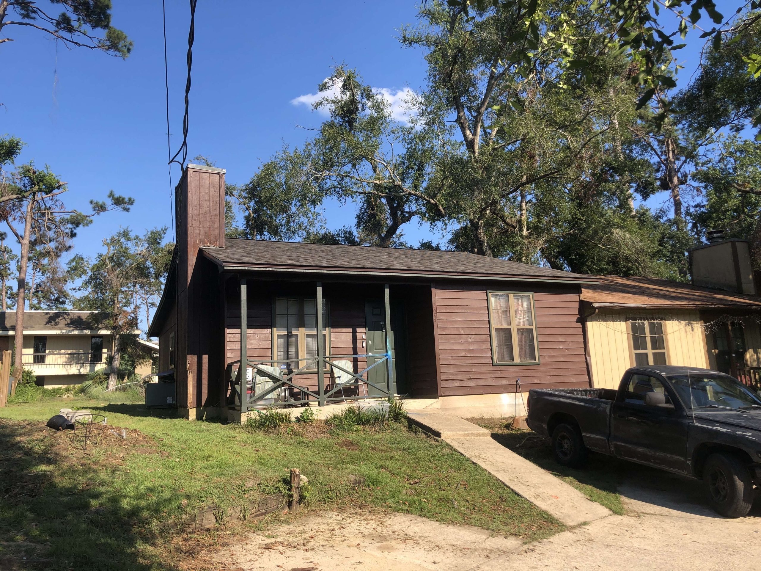 Single-story brown wooden house with a covered front porch, sloped driveway, and black pickup truck parked in front, surrounded by trees on a sunny day.