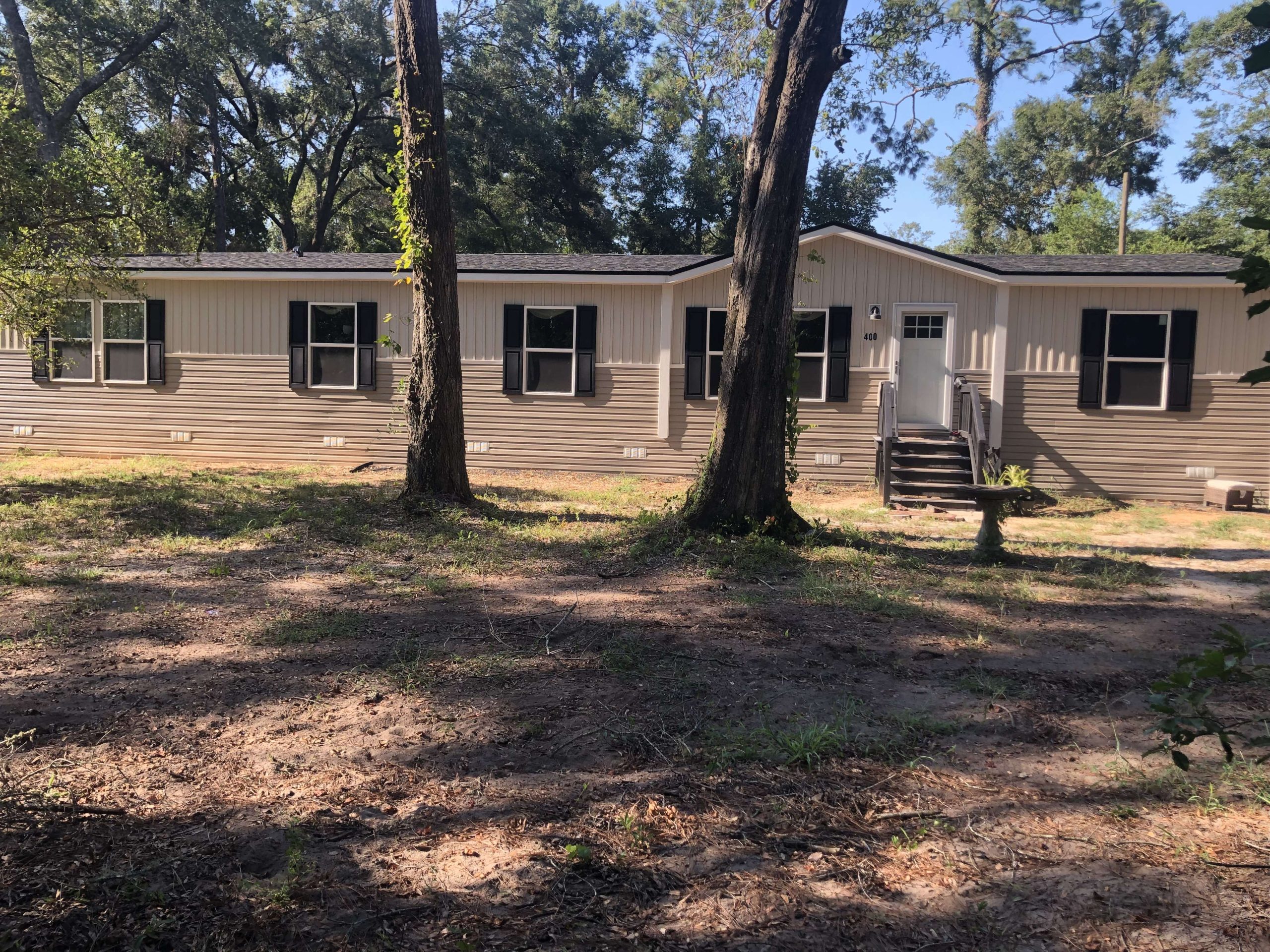 A beige double-wide mobile home with dark trim sits in a shaded yard with several tall trees and patchy grass.