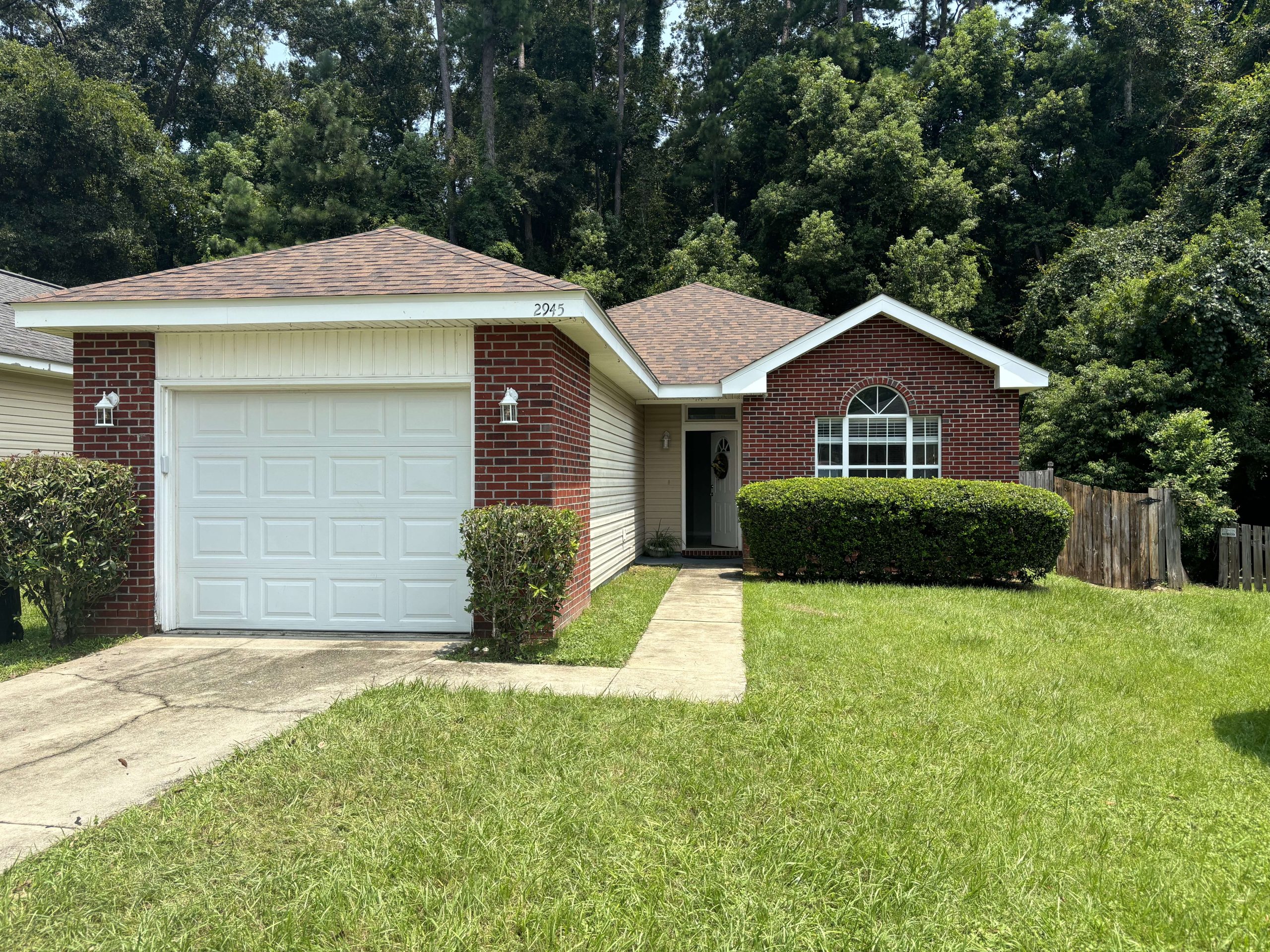 Single-story brick house with a white garage door, front lawn, bushes, and a walkway leading to the entrance. Trees and a wooden fence are in the background.