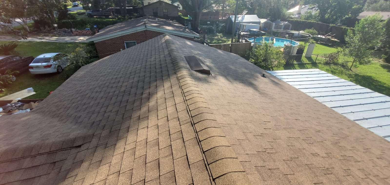 A view of a house roof with brown asphalt shingles, showing a rooftop vent; backyard with trees, a pool, and a parked car are visible.