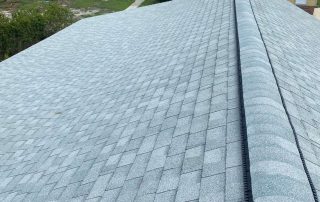 View from the peak of a house roof with new gray asphalt shingles and ridge vent installed, overlooking a yard, driveway, and trees in the background.