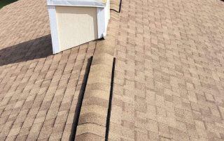 A house roof with brown shingles, a ridge vent running along the peak, and a white chimney pipe surrounded by trees and nearby rooftops.