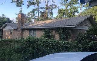 Single-story brick house with a gabled roof, chimney, and overgrown bushes in the front yard; trees and blue sky in the background.