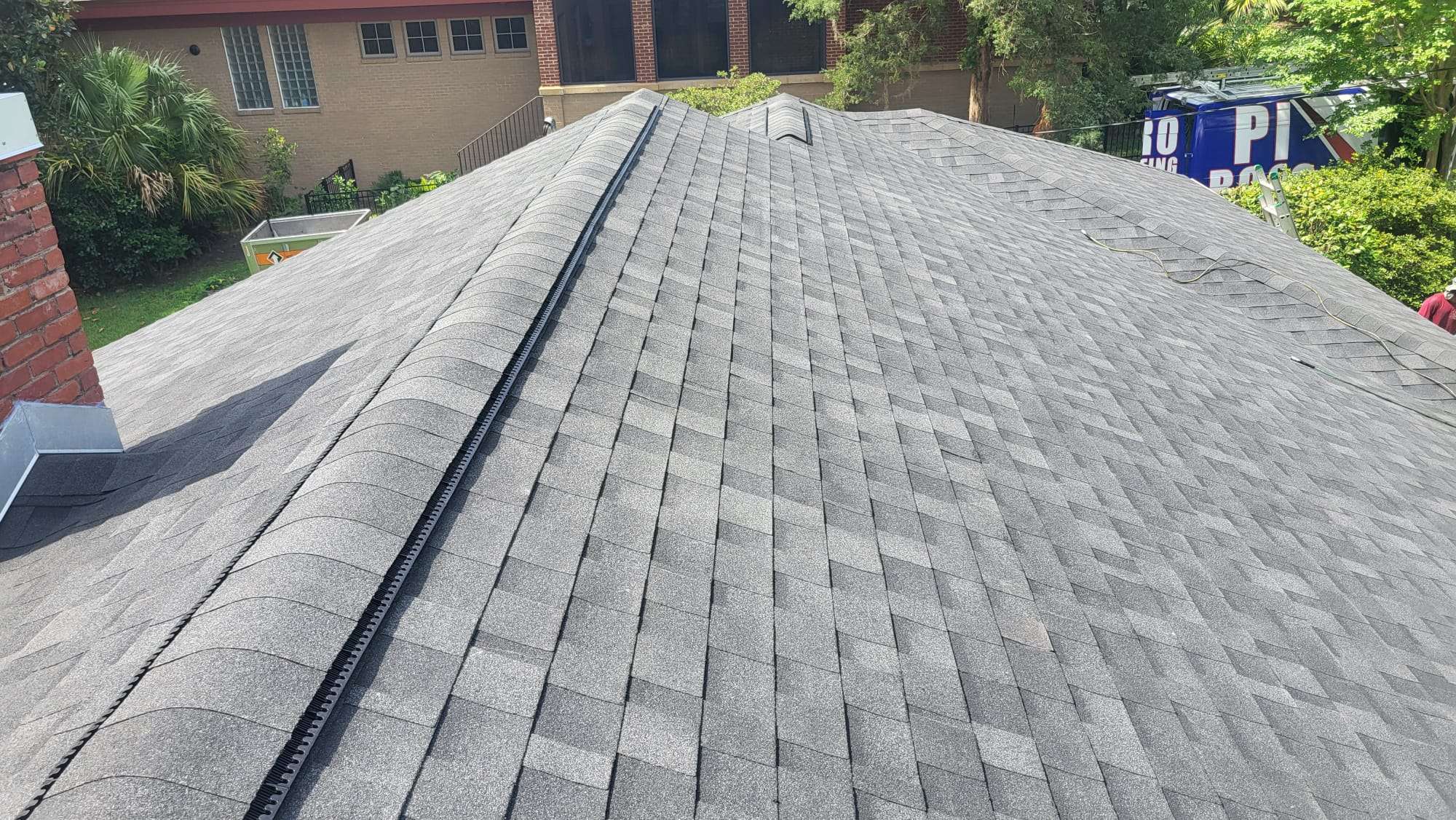 A house roof with gray asphalt shingles and a ridge vent, surrounded by trees and neighboring buildings.