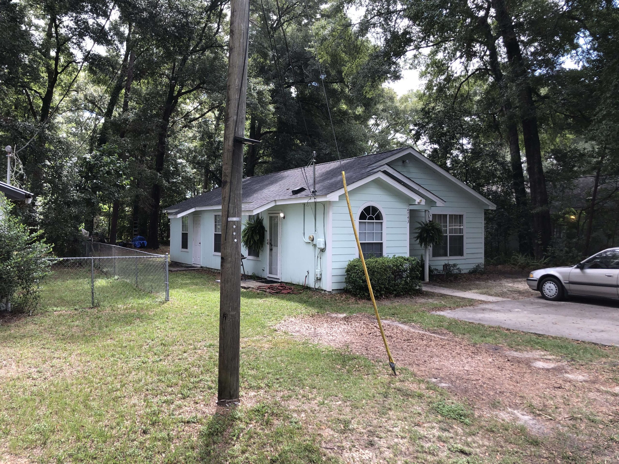 Single-story light blue house with white trim, surrounded by trees, a chain-link fence, and a driveway with a parked car. A utility pole stands in the front yard.