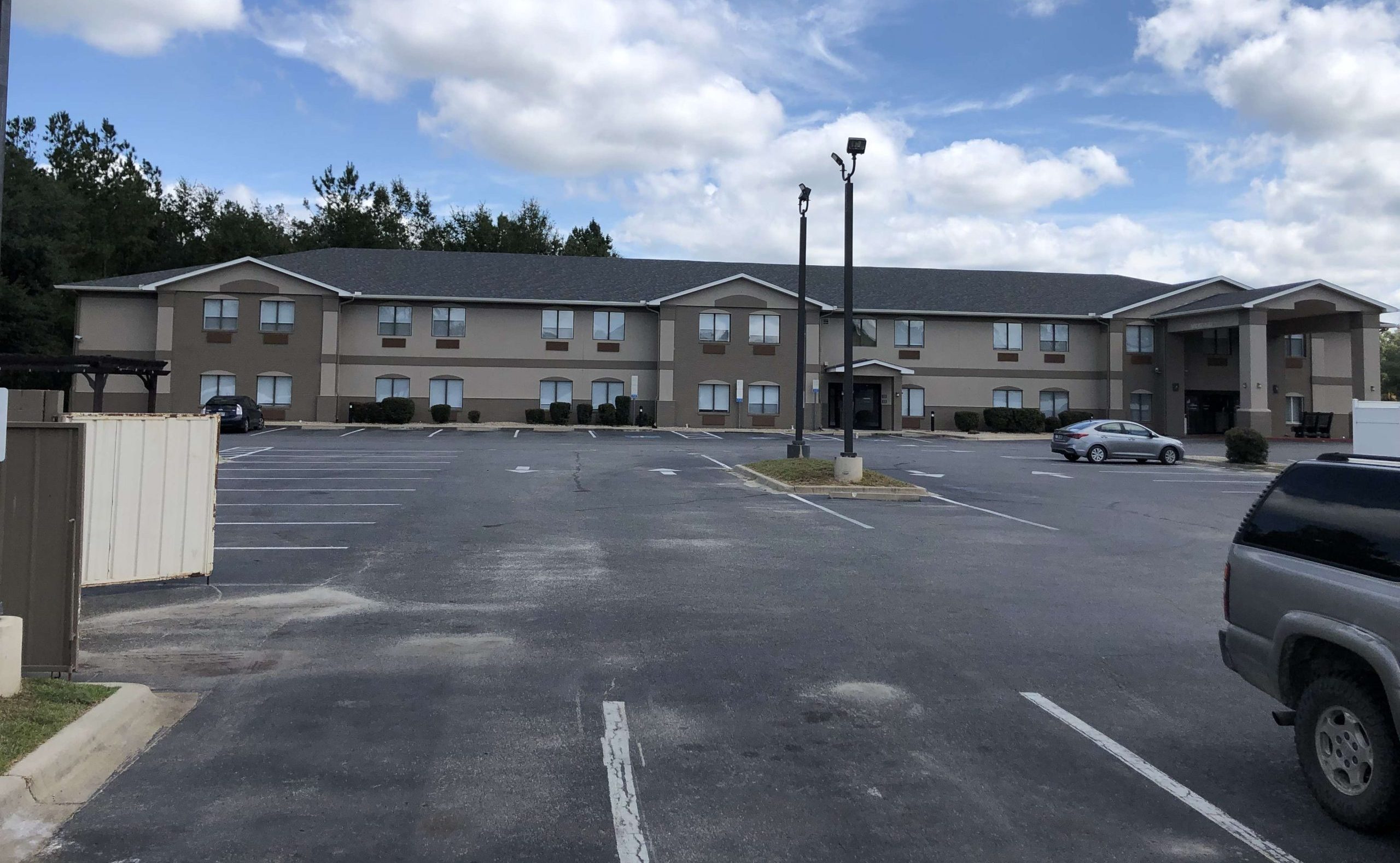 Two-story motel building with beige siding, several windows, and a mostly empty parking lot under a partly cloudy sky.