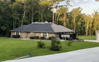 Single-story brick house with roofing materials stacked on the roof, a black roofing contractor trailer parked in the driveway, and trees in the background.