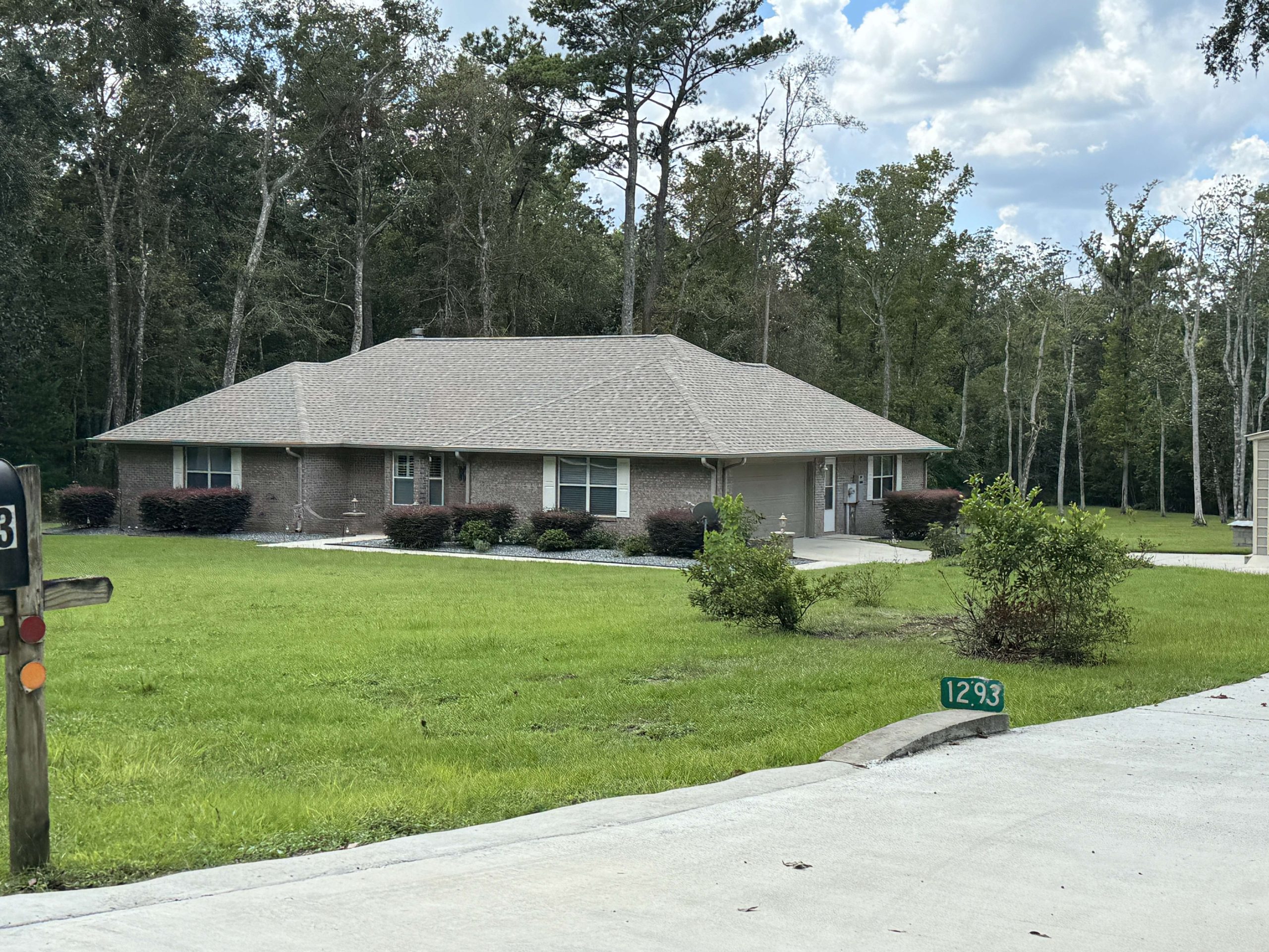 A single-story brick house with a gray roof sits on a large green lawn, surrounded by trees. A driveway and green address marker labeled 1293 are visible in the foreground.
