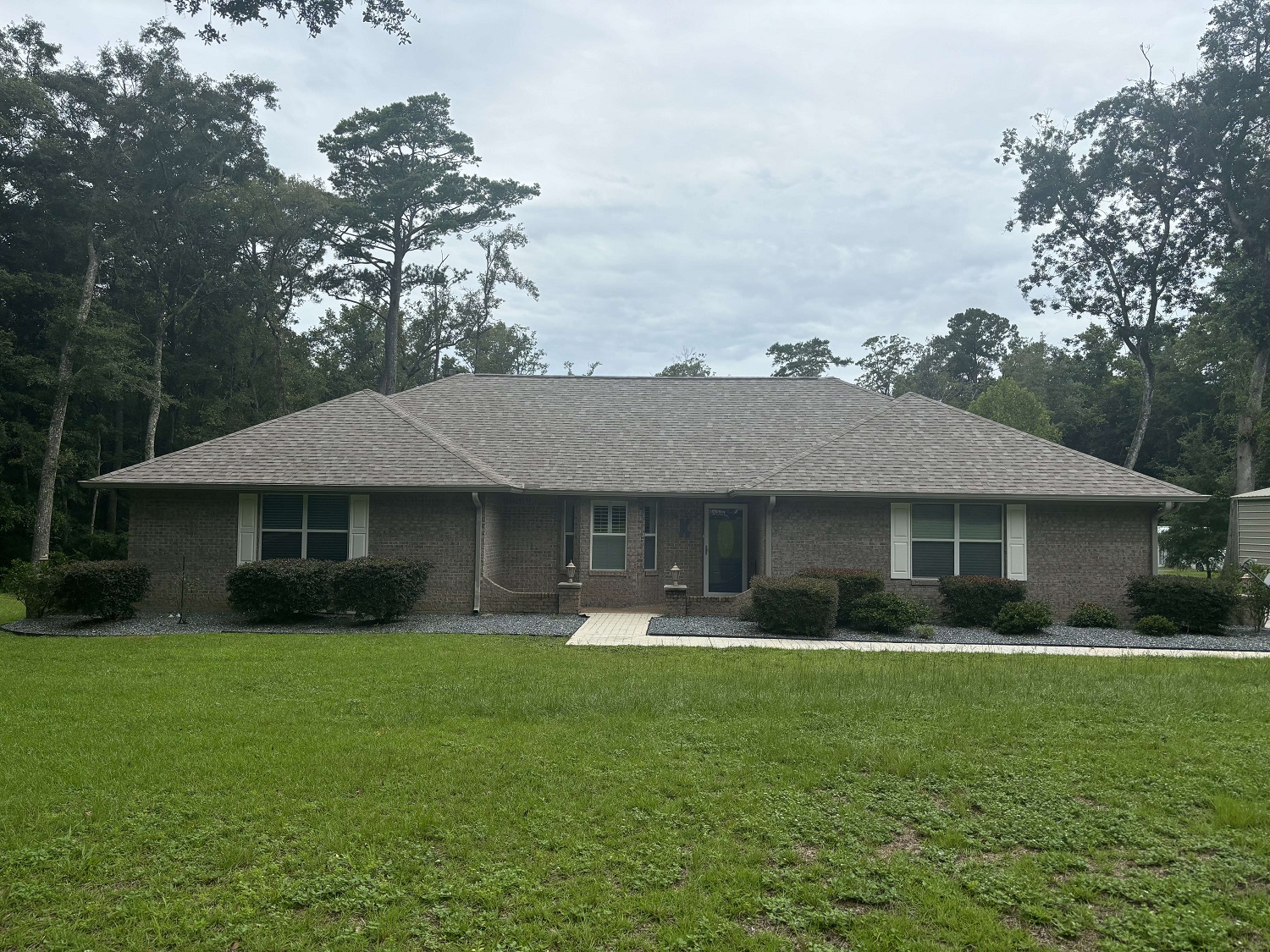 Single-story brick house with a hipped roof, central walkway, and surrounded by grass and trees under a cloudy sky.