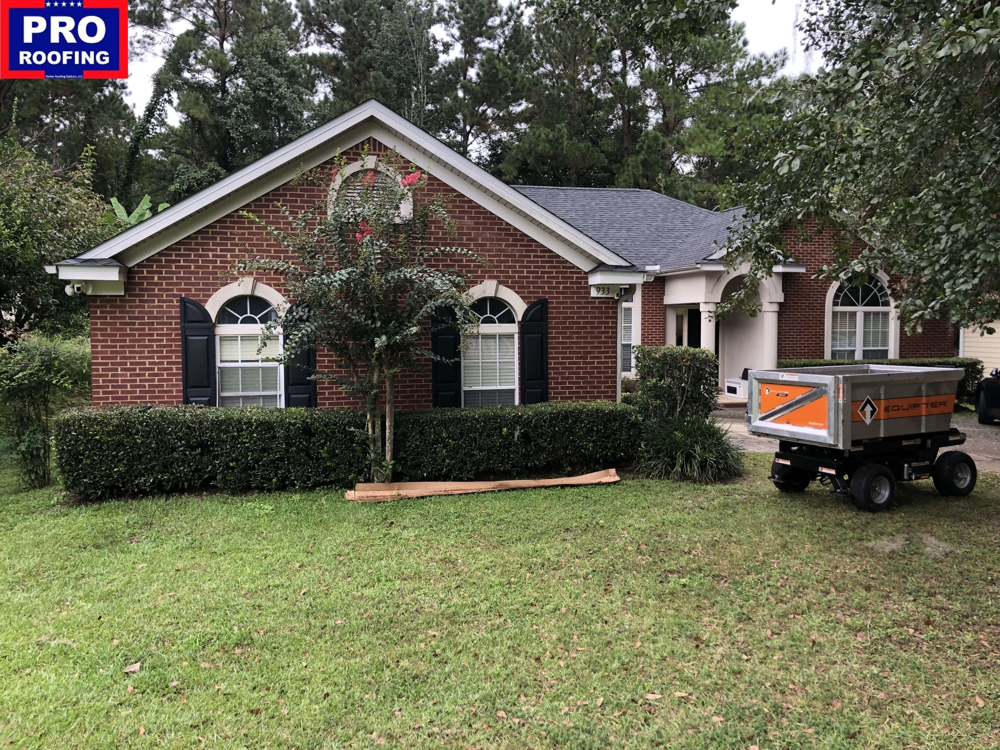 A brick house with a gray roof, arched windows, and a Pro Roofing sign in the corner. A small trailer is parked on the lawn near the driveway.