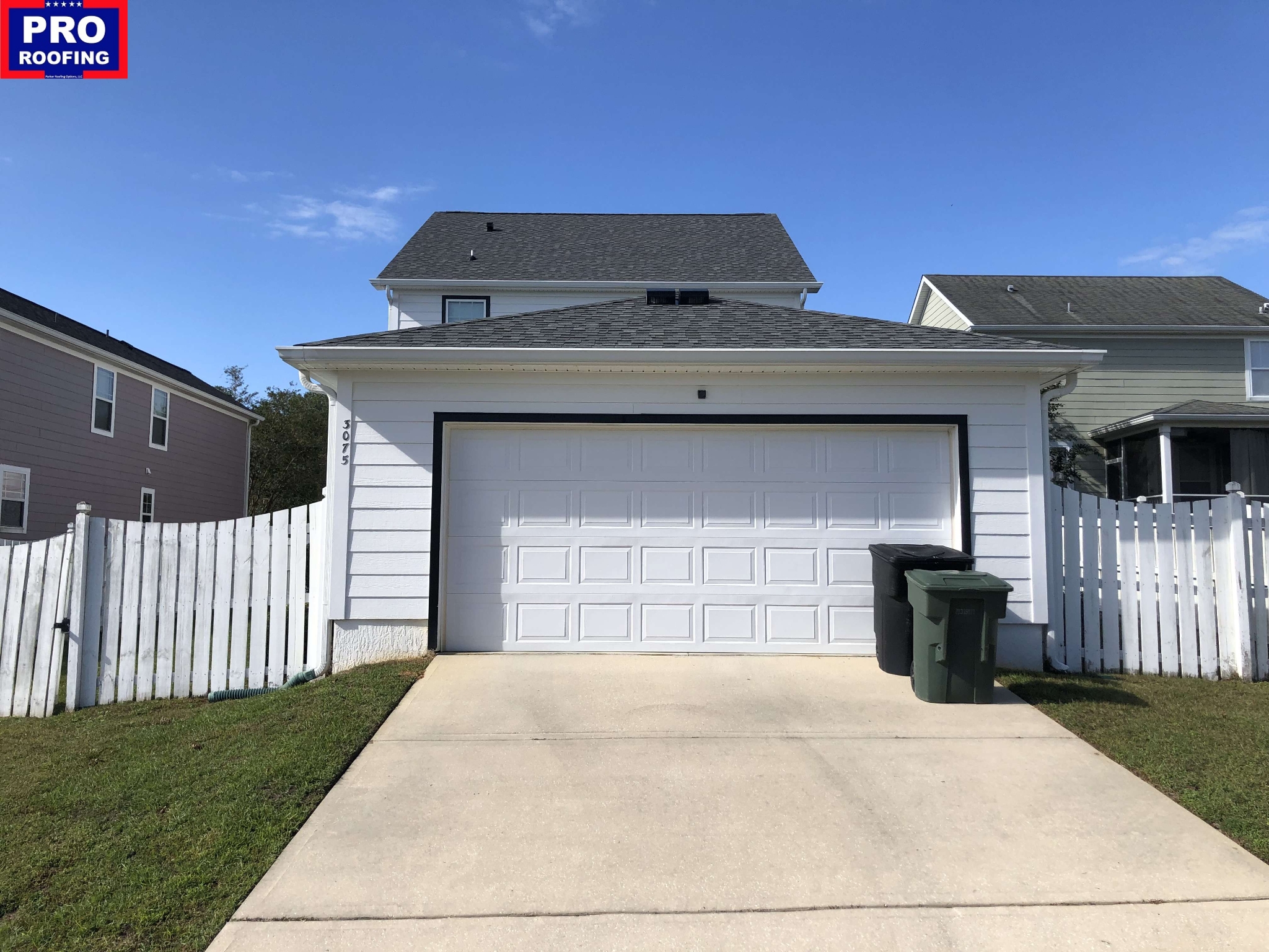 A white two-car garage with a driveway, two green trash bins, and a white picket fence; a PRO Roofing logo is in the images top left corner.