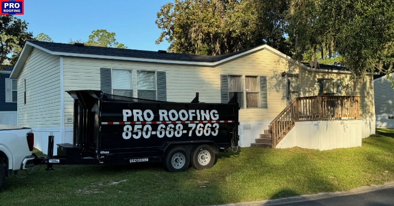 A single-story manufactured home with beige siding and a black dumpster labeled PRO ROOFING 850-668-7663 parked in front. The home has a small front porch and is surrounded by trees.