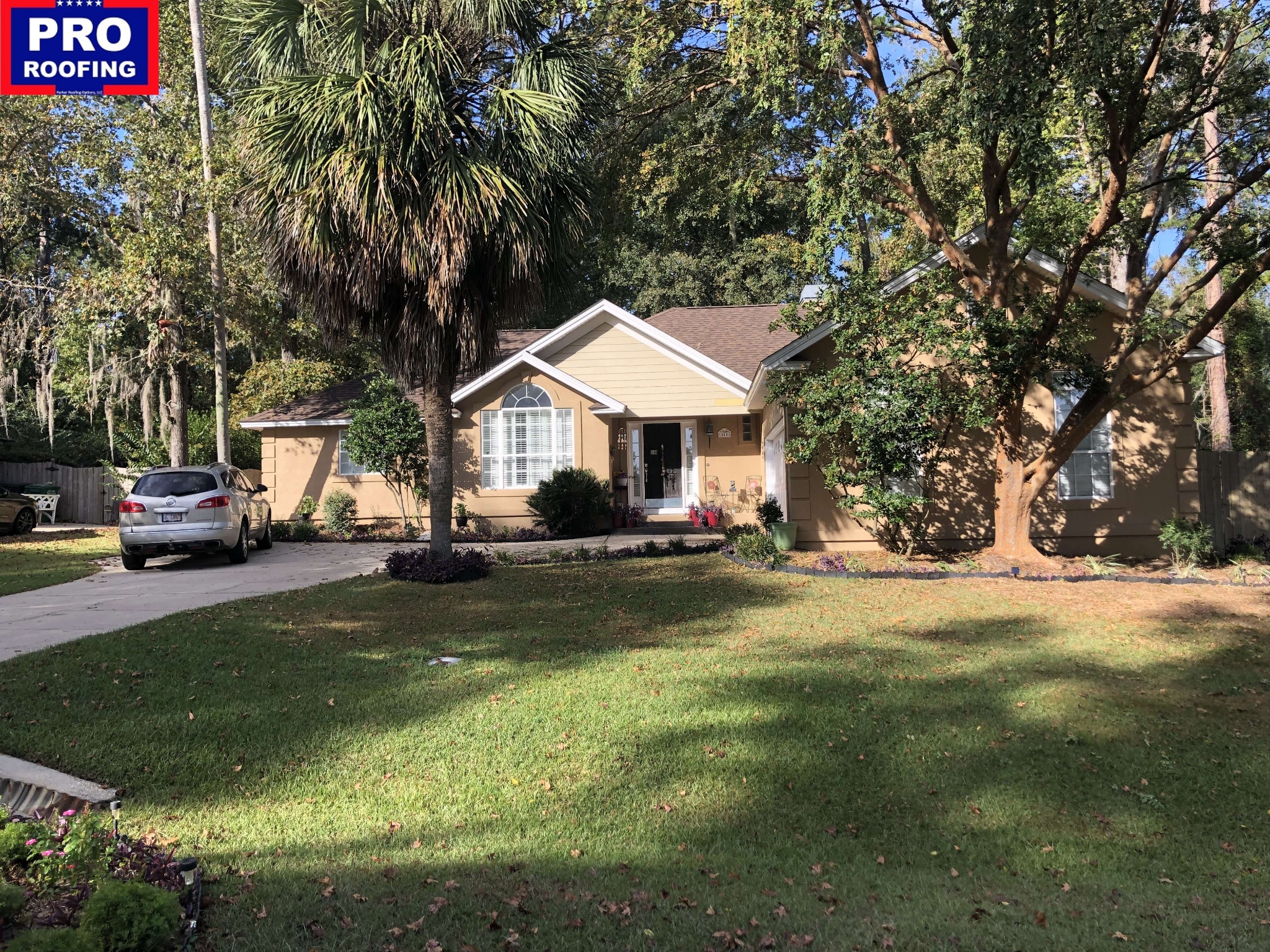 A tan single-story house with a brown roof, a parked car in the driveway, and trees and lawn in the front yard. A Pro Roofing logo is in the top left corner.