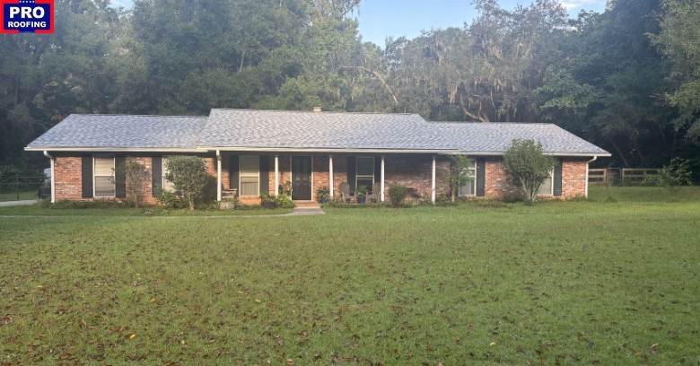 Single-story brick house with a gray shingle roof, white pillars, and surrounded by grass and trees. Pro Roofing logo appears in the top left corner.
