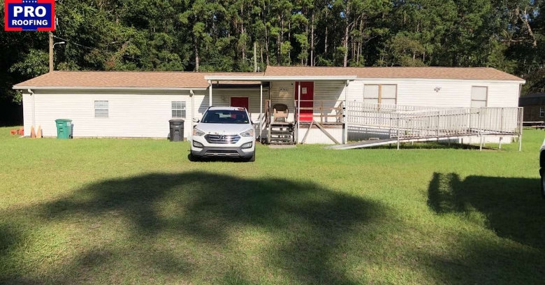 A white single-story house with a brown roof, wheelchair ramp, and a white SUV parked in front, set on a grassy lawn with trees in the background. Pro Roofing logo in the top left corner.