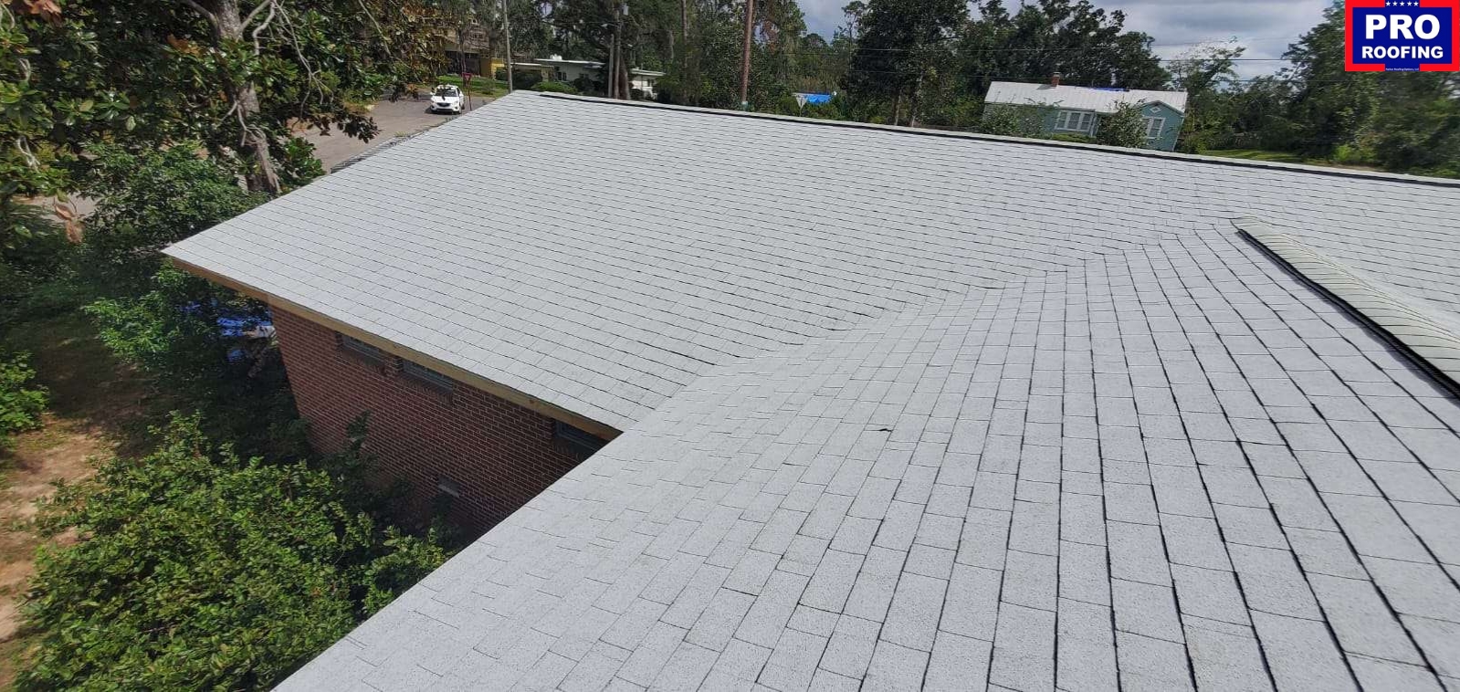 A light gray shingled roof on a brick building, surrounded by trees, with a PRO ROOFING sign in the upper right corner.