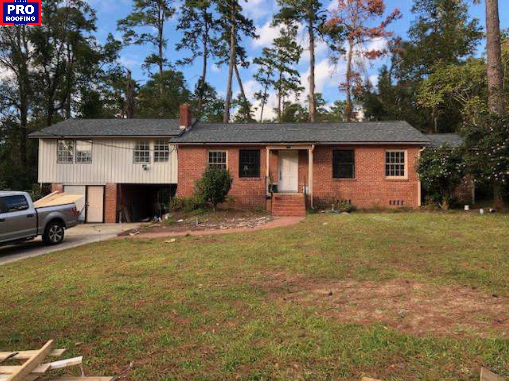 A split-level brick and siding house with a front porch, a driveway, a parked truck, and a yard with sparse grass and surrounding tall trees.