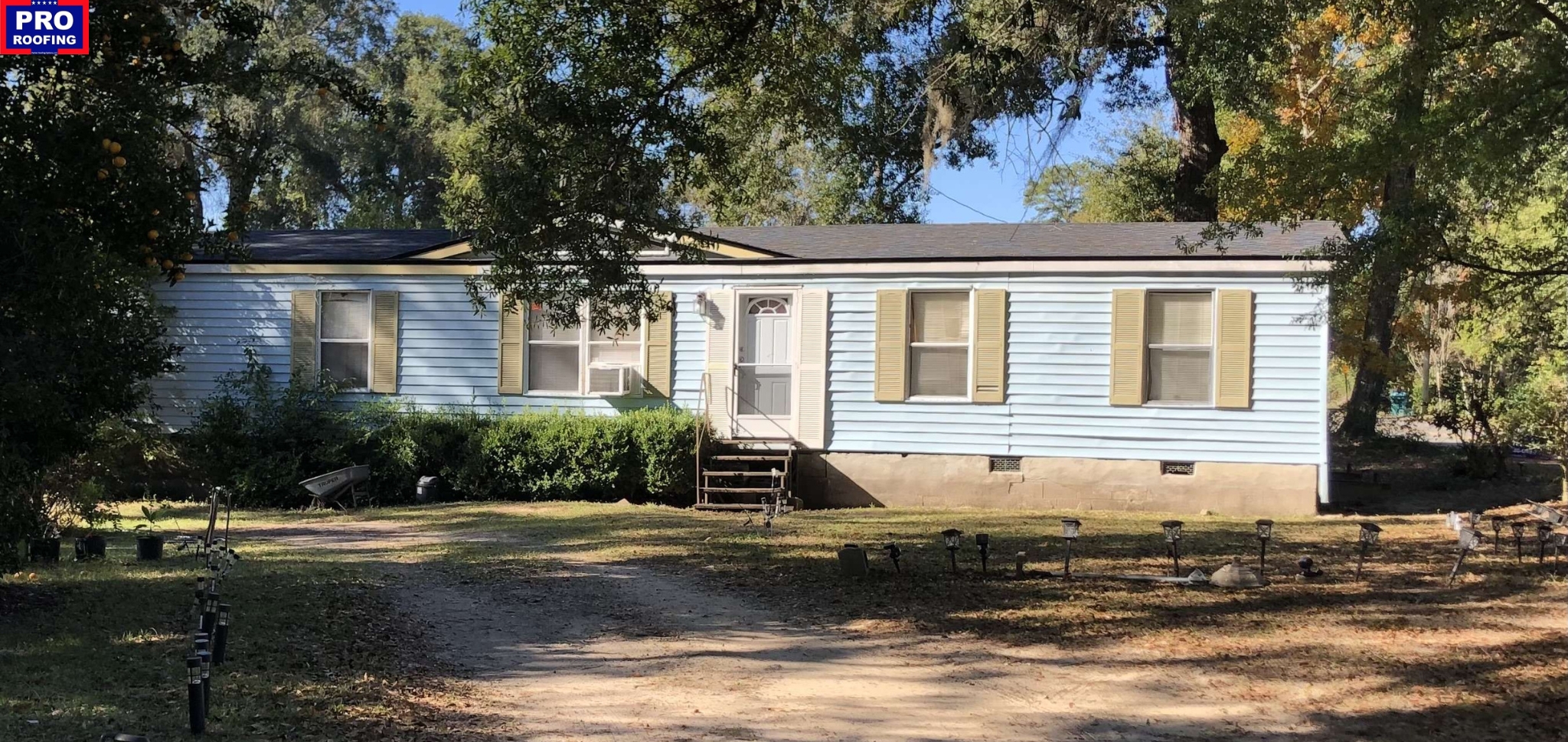 Single-story light blue manufactured home with tan shutters, a small front porch, and a yard with scattered trees and a dirt driveway; PRO Roofing sign in top left corner.
