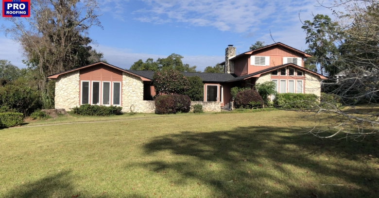 Two-story house with stone and wood exterior, multiple large windows, and a wide front lawn; Pro Roofing logo in the top left corner.