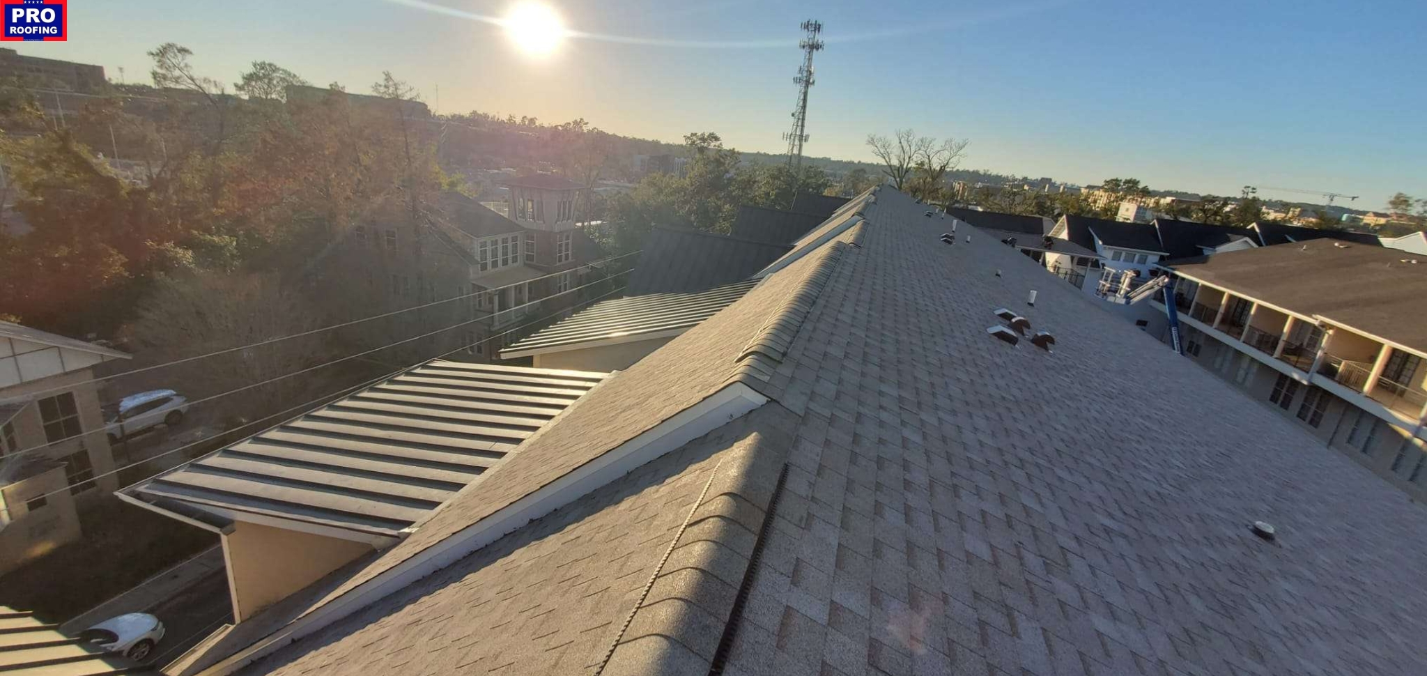 View of residential rooftops under a clear sky at sunset, with trees and buildings in the background and a Pro Roofing sign in the top left corner.