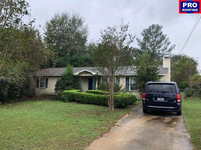 Single-story house with a gray roof, front bushes, and a black vehicle parked on a concrete driveway, with a Pro Roofing sign in the top right corner.