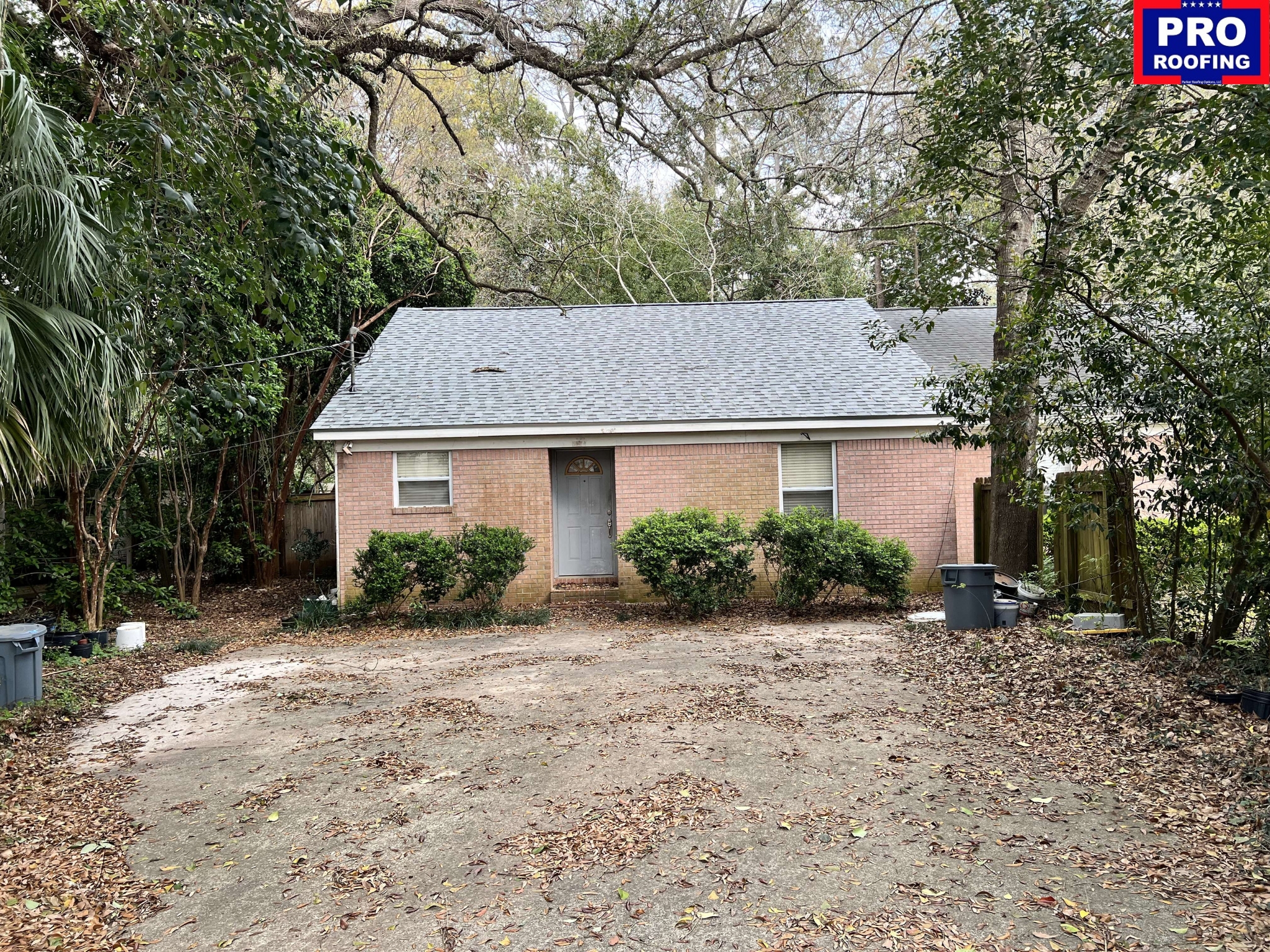 Single-story brick house with a gray shingle roof, surrounded by trees and bushes, with a PRO ROOFING sign in the top right corner.